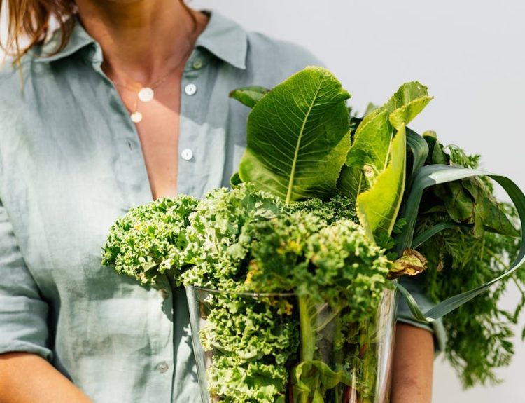 A woman in a light shirt carrying a basket of fresh greens, symbolizing healthy eating and lifestyle.