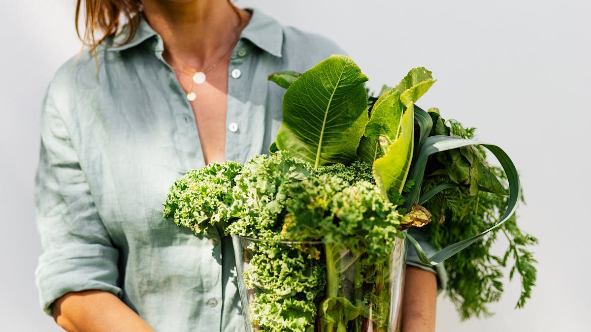 A woman in a light shirt carrying a basket of fresh greens, symbolizing healthy eating and lifestyle.